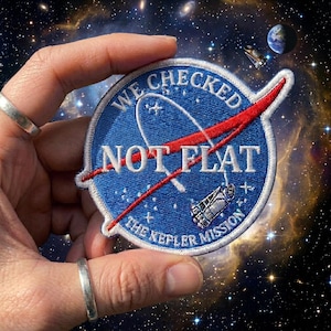 May include: A blue embroidered patch with the NASA logo and the text "WE CHECKED NOT FLAT THE KEPLER MISSION." The patch has a red swoosh and a white border, set against a space-themed background.