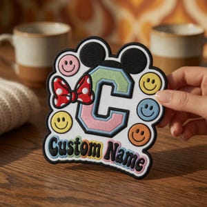 Könnte beinhalten: Ein weißer Stickaufnäher mit einem großen Buchstaben C in Pastellgrün und Rosa, gekrönt von schwarzen Mausohren und einer roten Schleife mit weißen Punkten. Der Aufnäher ist von bunten Smileys und dem schwarzen Schriftzug Custom Name umgeben.