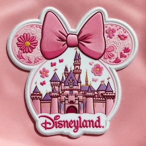 May include: Pink embroidered patch in the shape of Minnie Mouse's head, featuring a pink bow, flowers, and a castle. The word "Disneyland" is embroidered in pink script at the bottom. The patch is on a pink background.