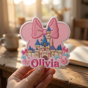 May include: A pink embroidered patch shaped like a mouse head with a large bow, featuring a detailed castle illustration in blue, white, and tan. The name Olivia is stitched in bold pink letters at the bottom, surrounded by small pink and blue flowers.