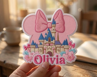 May include: A pink embroidered patch shaped like a mouse head with a large bow, featuring a detailed castle illustration in blue, white, and tan. The name Olivia is stitched in bold pink letters at the bottom, surrounded by small pink and blue flowers.