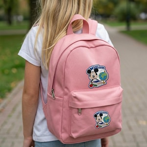 May include: A pink backpack with a front pocket and a top handle. The backpack features two patches of Mickey Mouse with a globe and books, with the name "William" on the patch. The backpack is being worn by a person.