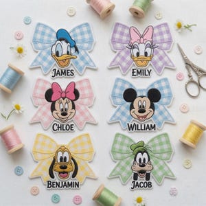 May include: Six embroidered patches featuring Disney characters on gingham bows with names James, Emily, Chloe, William, Benjamin, and Jacob. The patches are arranged on a white surface with scattered sewing spools, buttons, and small white flowers.