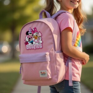 May include: Pink backpack with a front pocket and lavender accents. Features a graphic of Minnie Mouse and Daisy Duck with the word "Besties". Includes the text "Oh Joy! Section" on the front.