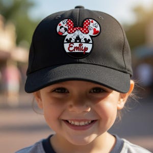 May include: Black baseball cap with a Minnie Mouse design, a red bow, and heart details. The cap displays the name "Emily" in white and red. The cap is worn by a smiling child.