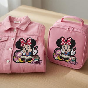 Pu&ograve; includere: Una giacca di jeans rosa e un portapranzo abbinato con un motivo di Minnie Mouse. Il motivo mostra Minnie Mouse con un fiocco rosso a pois, che tiene una penna e fa il segno della pace. Il nome "Emily" &egrave; stampato sotto.