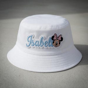 May include: White bucket hat with a light blue embroidered patch that reads "Isabell" with a Minnie Mouse graphic. The hat is made of a soft material and has a rounded crown and a downward-sloping brim.