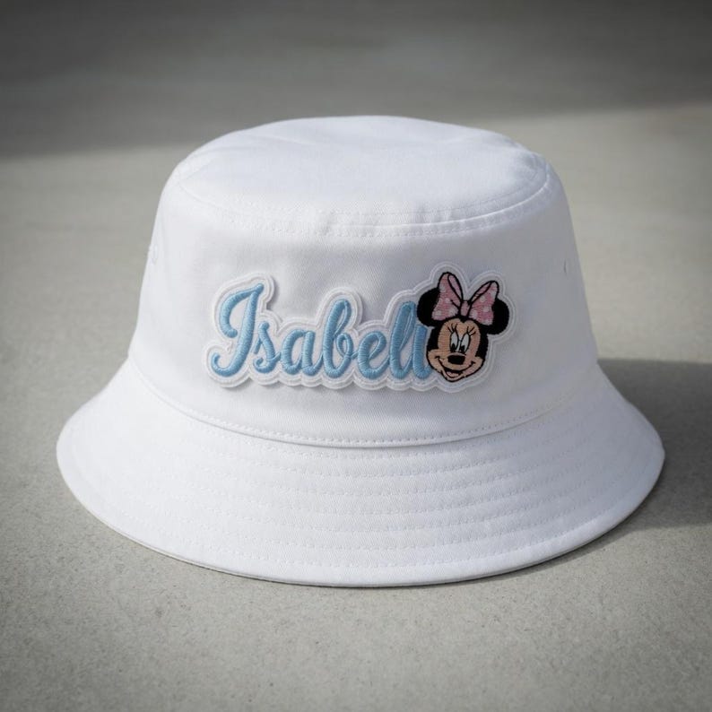 May include: White bucket hat with a light blue embroidered patch that reads "Isabell" with a Minnie Mouse graphic. The hat is made of a soft material and has a rounded crown and a downward-sloping brim.