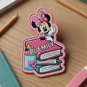 May include: A pink embroidered patch featuring Minnie Mouse with a polka dot bow, sitting atop a stack of books. The name "EMILY" is printed on the top book. A pencil holder with pencils and pens is next to the books.
