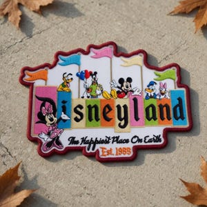 May include: Embroidered patch featuring the Disneyland logo with Mickey Mouse, Minnie Mouse, Donald Duck, and Pluto. The patch has a red border and includes the text "The Happiest Place On Earth" and "Est. 1955".