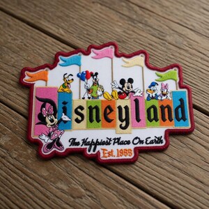 May include: Embroidered Disneyland patch featuring Mickey Mouse, Minnie Mouse, Donald Duck, and Pluto. The patch has a white background with colorful letters and flags, and the text "The Happiest Place On Earth" and "Est. 1955".