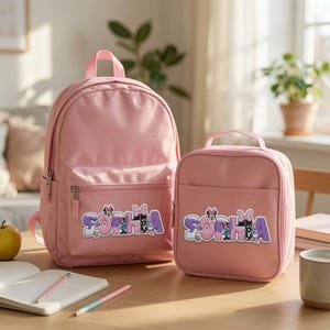 May include: A light pink backpack and a matching lunch bag, both featuring the name SOPHIA in large, purple and white block letters decorated with small cartoon character graphics and polka-dot bow patterns on the front pockets.