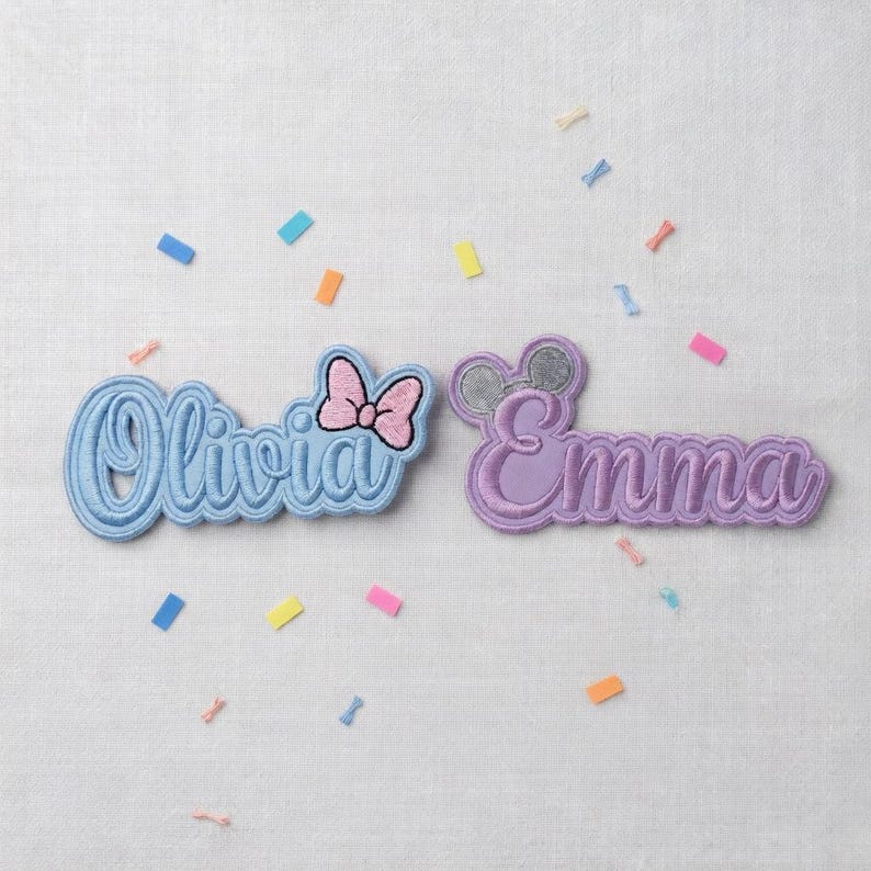 May include: Two embroidered name patches, "Olivia" in light blue with a pink bow, and "Emma" in purple with mouse ears. Colorful confetti is scattered around the patches on a white background.