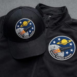 Può includere: Cappellino da baseball e giacca neri con una toppa rotonda a tema spaziale. La toppa presenta un pianeta giallo, un razzo, stelle e la scritta "Astral Pioneer Division".