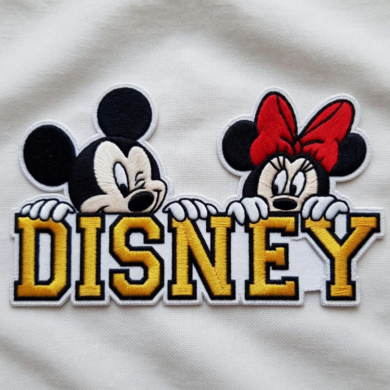 Puede incluir: Parche bordado de Disney con Mickey y Minnie Mouse sobre la palabra "DISNEY" en oro y blanco. Mickey gui&ntilde;a un ojo y Minnie lleva un lazo rojo. El parche est&aacute; sobre un fondo blanco.