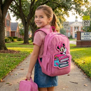 May include: A pink backpack with a Minnie Mouse patch, a matching lunch bag, and a girl wearing a red and white checkered shirt and denim skirt. The backpack has a patch with the name "Emily" on it.