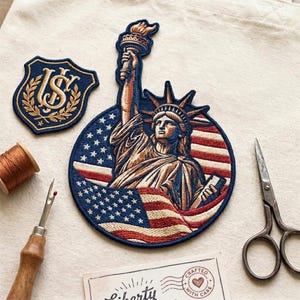 Könnte beinhalten: Gestickte Aufnäher mit der Freiheitsstatue und einem Schild mit den Buchstaben "US" in Gold. Der Aufnäher der Freiheitsstatue ist kreisförmig, mit blauem Hintergrund und rot-weißen Streifen. Eine Garnrolle, ein Nahttrenner, eine Schere und eine Karte mit "Liberty" sind ebenfalls vorhanden.