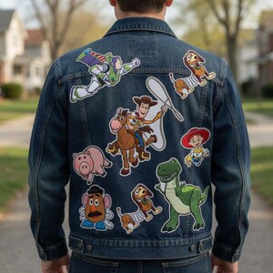K&ouml;nnte beinhalten: Eine dunkelblaue Jeansjacke mit mehreren gestickten Aufn&auml;hern von Zeichentrickfiguren, darunter ein Weltraum-Ranger, ein Cowboy auf einem Pferd mit Lasso, ein Cowgirl, ein gr&uuml;ner Dinosaurier, ein Schwein, eine Kartoffelfigur und zwei Federhunde.