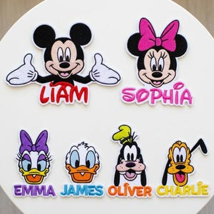 May include: Embroidered patches featuring Disney characters Mickey Mouse, Minnie Mouse, Daisy Duck, Donald Duck, Goofy, and Pluto. Each character has a name embroidered below: Liam, Sophia, Emma, James, Oliver, and Charlie. The patches are colorful and detailed.