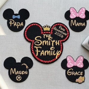 Könnte beinhalten: Schwarze Mickey Mouse-förmige Aufnäher mit gestickten Namen und Details. Einer trägt die Aufschrift "Papa" mit einer blauen Schleife. Ein anderer trägt die Aufschrift "Mama" mit einer rosa Schleife. Ein großer Aufnäher trägt die Aufschrift "The Smith Family Park Adventure 2026".