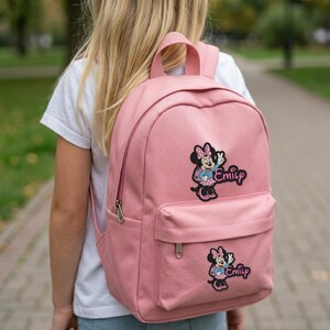 Puede incluir: Una mochila rosa con un bolsillo frontal y un asa superior. La mochila presenta dos parches de Minnie Mouse con el nombre "Emily" bordado debajo. La mochila est&aacute; hecha de un material duradero y tiene correas ajustables.