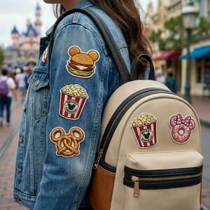 May include: A light-wash denim jacket and a beige backpack adorned with colorful patches. The jacket features patches of a burger, popcorn, and a pretzel. The backpack has popcorn and a donut patches.