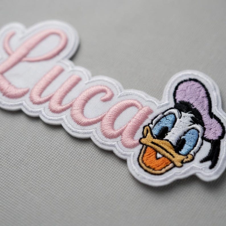 May include: Embroidered patch featuring the name "Luca" in pink script, with a white scalloped border. The patch includes a cartoon image of Donald Duck. The patch is designed for clothing or accessories.