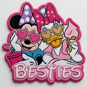 May include: Embroidered patch featuring Minnie Mouse and Daisy Duck wearing heart-shaped sunglasses. Minnie holds a drink, Daisy holds an ice cream cone. The word "BESTIES" is in white, outlined in pink.