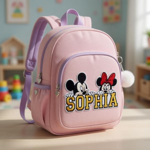 Op de afbeelding: Een roze rugzak met paarse accenten, met Mickey en Minnie Mouse. De naam "SOPHIA" is geborduurd in geel en wit. Inclusief een witte pompom-bedel en hartvormige ritstrekkers. Ideaal voor school of reizen.