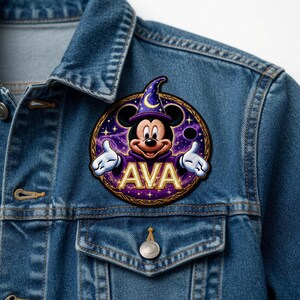 May include: A circular patch featuring Mickey Mouse in a wizard's hat and robe, with the name "AVA" in gold lettering. The patch has a purple and gold color scheme, with stars and a crescent moon design. It is affixed to a blue denim jacket.