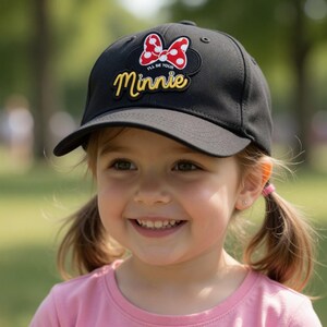 May include: Black baseball cap with a curved peak. The cap features a Minnie Mouse design with a red and white polka dot bow and the text "I'll Be Your Minnie" in yellow. The cap is worn by a young girl.