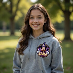 May include: A gray hooded sweatshirt featuring a circular graphic of Mickey Mouse in a wizard hat, with the name "SARA" in gold lettering. The graphic is set against a purple and gold starry background. The person is smiling.