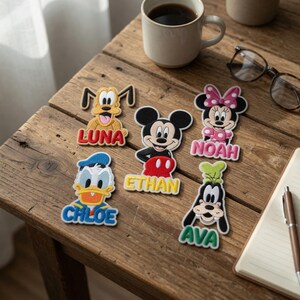 May include: A collection of colorful Disney character patches with names, including Pluto, Mickey Mouse, Minnie Mouse, Donald Duck, and Goofy. Each patch features a different character with a personalized name in a contrasting color. The patches are on a wooden surface.