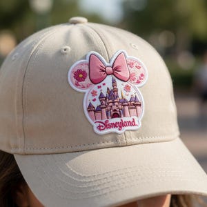 May include: Beige baseball cap with a pink Disneyland patch. The patch is shaped like a mouse head, with a pink bow, castle, and the word "Disneyland". The cap has a curved peak and a button on the crown.