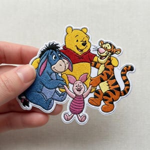 Puede incluir: Parche bordado con Winnie the Pooh, Tigger, Eeyore y Piglet. Winnie the Pooh es amarillo, con una camisa roja. Tigger es naranja con rayas negras. Eeyore es azul y Piglet es rosa.