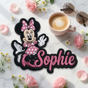 May include: An embroidered patch featuring the cartoon character Minnie Mouse wearing a pink polka-dot dress and bow, positioned next to the name Sophie written in a pink cursive font with a thick black outline on a white fabric backing.