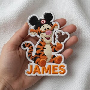 May include: Embroidered patch featuring Tigger from Winnie the Pooh wearing a Mickey Mouse hat and holding a Mickey-shaped ice cream bar. The name "JAMES" is embroidered in orange on a white background.
