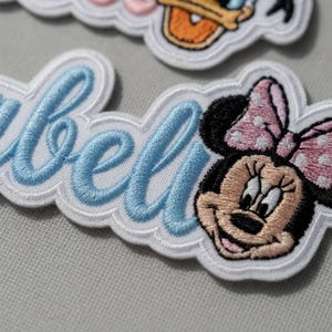 May include: Embroidered patch featuring the name "Isabel" in light blue script with a white border. A Minnie Mouse cartoon character is on the right, with a pink bow with white polka dots. The patches are on a light gray background.