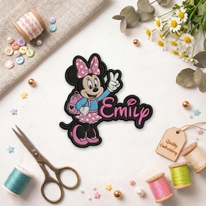 Puede incluir: Parche bordado con Minnie Mouse con un vestido y lazo de lunares rosas, haciendo el signo de la paz. El nombre "Emily" est&aacute; escrito en cursiva rosa. Una peque&ntilde;a etiqueta dice "Quality Embroidery".