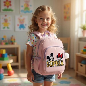Puede incluir: Una mochila rosa claro con un dise&ntilde;o de Mickey Mouse y Minnie Mouse, y el nombre "SOPHIA" en letras doradas. La mochila tiene un peque&ntilde;o bolsillo frontal, un asa superior y un pomp&oacute;n blanco. La ni&ntilde;a lleva una camiseta blanca con lunares de colores.
