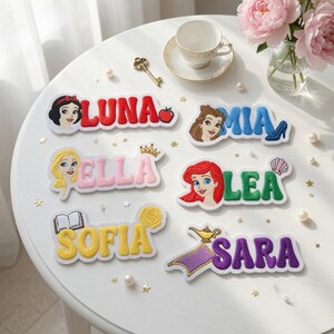 Peut inclure: Six écussons brodés avec des noms, des personnages de dessins animés et des icônes : LUNA avec une pomme, MIA avec un soulier bleu, ELLA avec une couronne, LEA avec un coquillage, SOFIA avec un livre et une rose jaune, SARA avec une lampe.