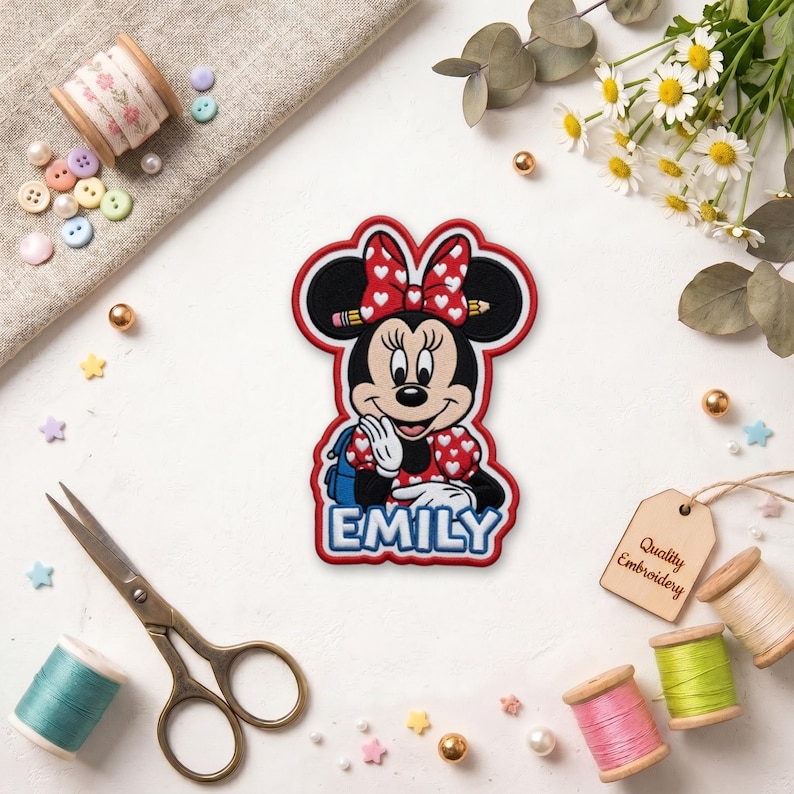 May include: Embroidered patch featuring Minnie Mouse with a red bow and dress with white hearts. The name "EMILY" is displayed in blue letters. The patch is surrounded by sewing supplies, including thread spools and scissors.
