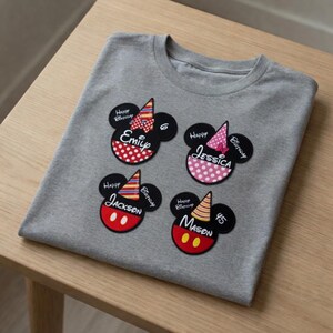 以下が含まれることがあります： グレーのTシャツに、ミッキーマウスをテーマにした4つの誕生日パッチが付いています。各パッチには名前、誕生日の帽子、そして「Happy Birthday」の文字が描かれています。パッチは赤、ピンク、黒のアクセントがあります。