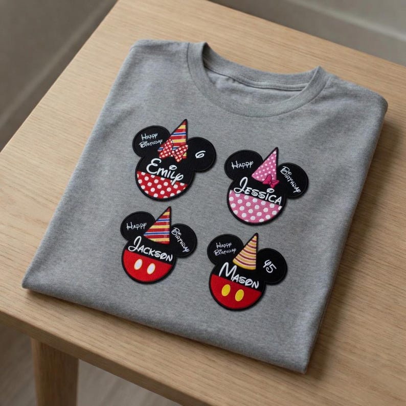 以下が含まれることがあります： グレーのTシャツに、ミッキーマウスをテーマにした4つの誕生日パッチが付いています。各パッチには名前、誕生日の帽子、そして「Happy Birthday」の文字が描かれています。パッチは赤、ピンク、黒のアクセントがあります。