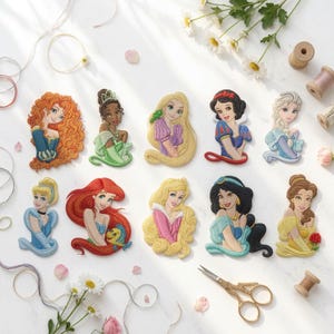 May include: A collection of ten embroidered patches featuring various animated female characters with distinct hair colors, clothing styles, and expressions, arranged in two rows on a white surface alongside sewing supplies and white flowers.