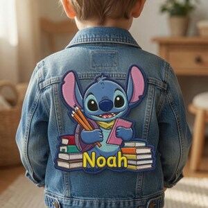 May include: A blue denim jacket featuring a large embroidered patch on the back. The patch depicts a blue alien character holding pencils and a pink book, surrounded by stacks of colorful books with the name Noah written in yellow letters.