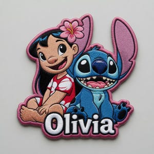 May include: An embroidered patch featuring a cartoon girl with black hair and a pink hibiscus flower, sitting next to a blue alien character with large ears. The name Olivia is stitched in white letters at the bottom, all outlined in pink thread.