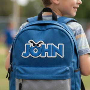 以下が含まれることがあります： グレーの底部分が付いた青いバックパック。「JOHN」という名前が白い文字で書かれており、その上に漫画の目のデザインがあります。黒いストラップとジッパーが付いています。