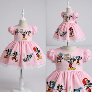 May include: Pink dress with short sleeves and a white lace trim. The dress features embroidered Mickey Mouse characters and birthday-themed designs. The dress is displayed on a mannequin.