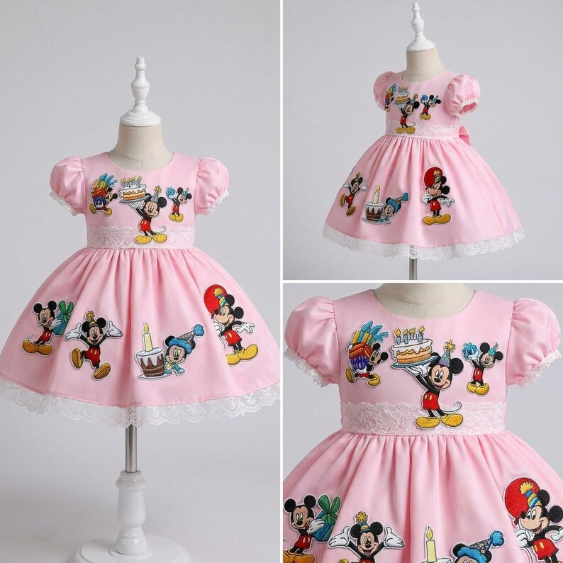 May include: Pink dress with short sleeves and a white lace trim. The dress features embroidered Mickey Mouse characters and birthday-themed designs. The dress is displayed on a mannequin.